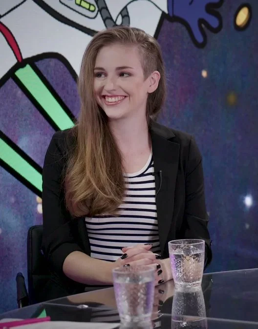 A photograph of Molly White sitting at a table in a video studio. She's
          a white woman with long brown hair that's shaved on one side, she has brown eyes, and she's wearing a black and
          white striped shirt and black blazer. There is a colorful cartoon background behind her. She's smiling and looking
          at someone out of frame.